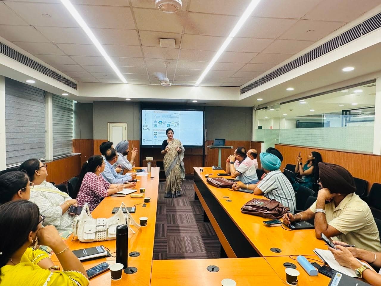 Teaching at the head of a seminar room, standing beside a projection screen while participants seated around a U-shaped table turn towards her.