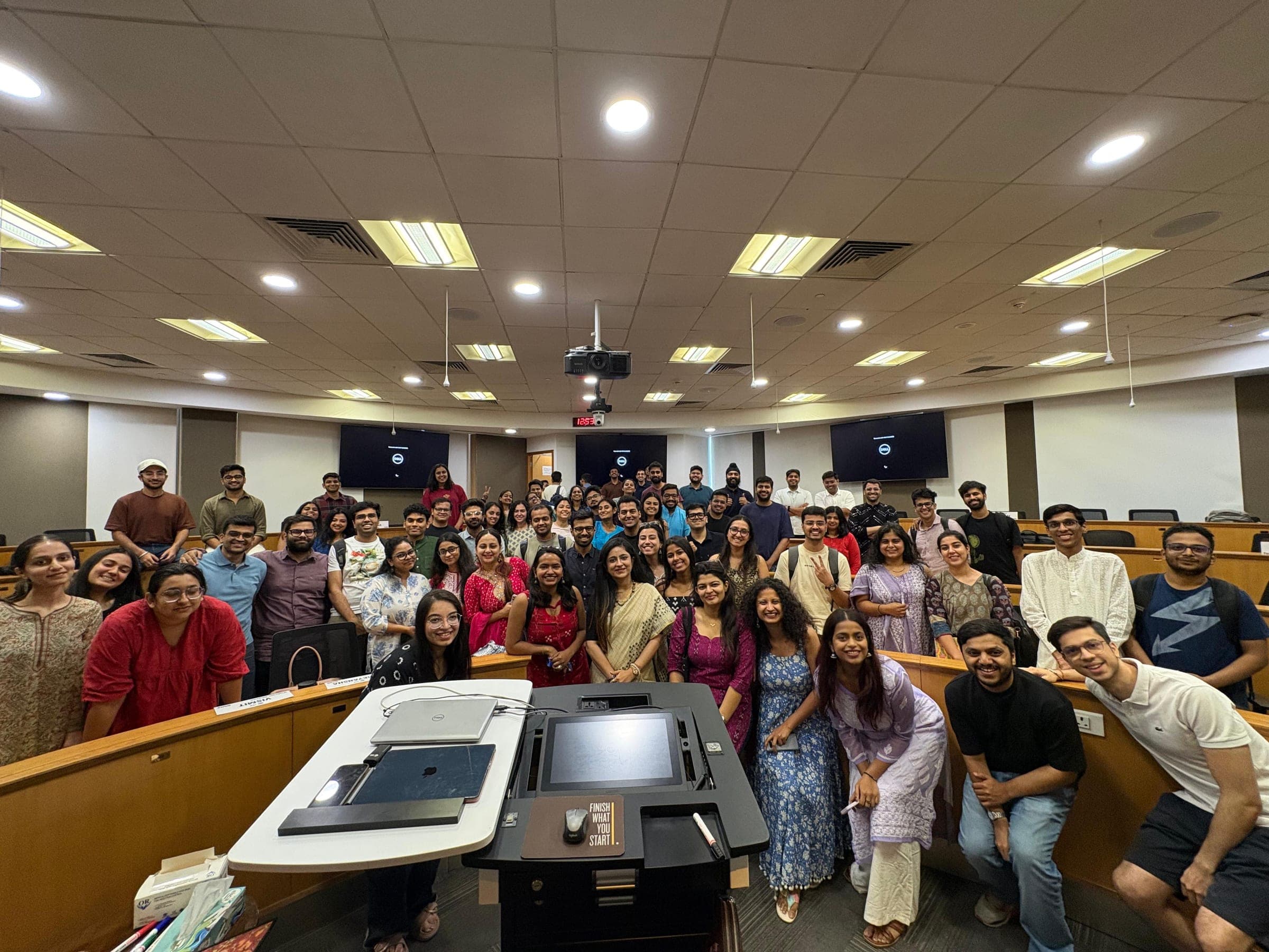 Standing with a full student cohort crowded together on the tiered steps of a lecture theatre, the lectern and its monitor in the foreground.