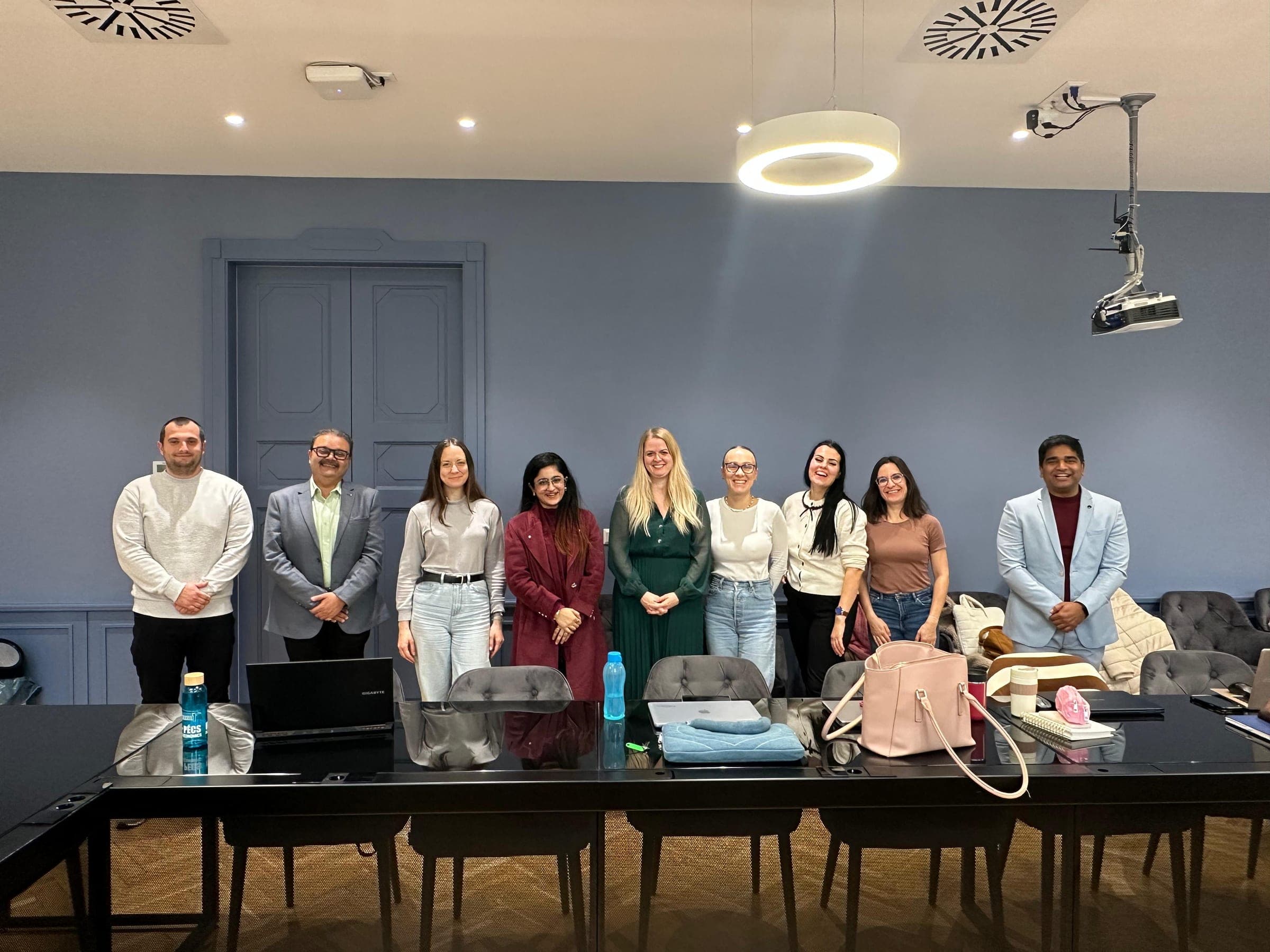 Standing in a line with an international group of students in a high-ceilinged European classroom, their laptops and bags on the desks in front of them.