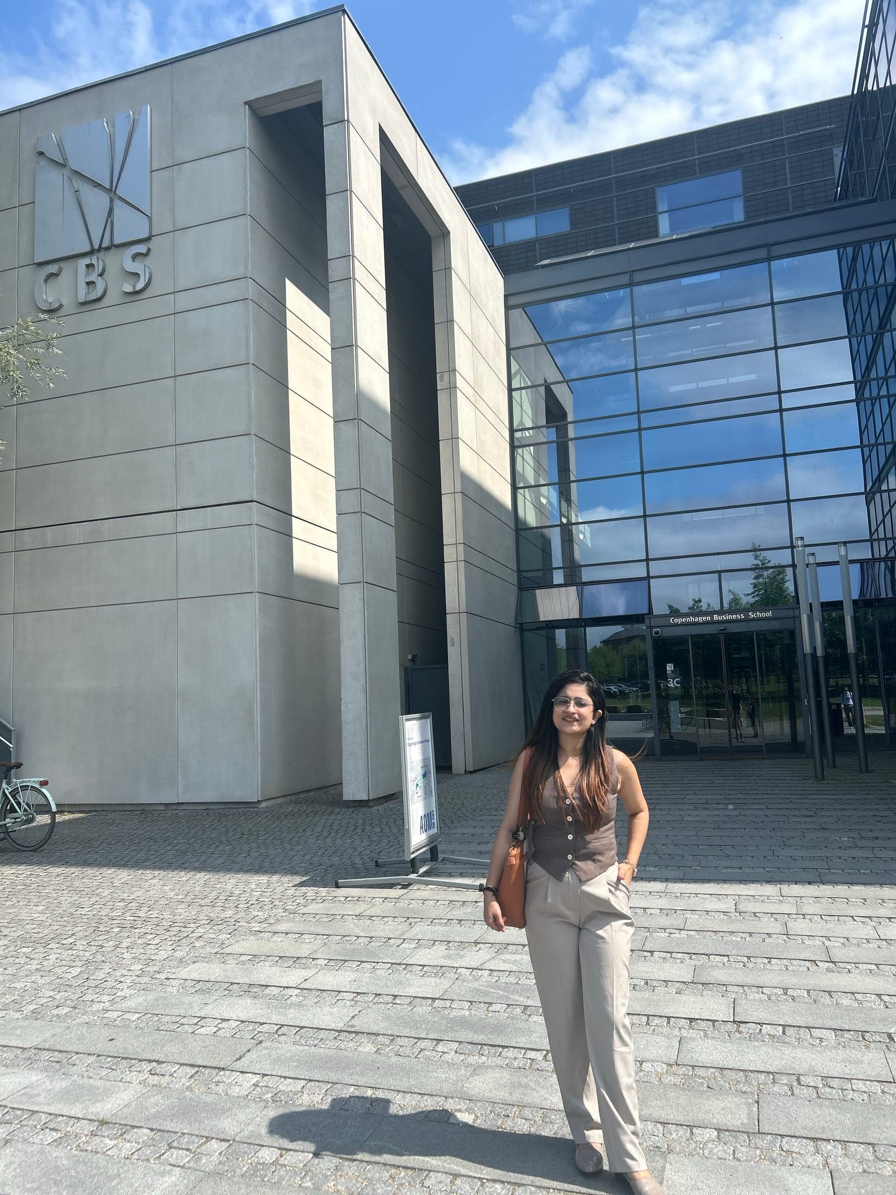 Walking down the front steps of Copenhagen Business School, the glass and concrete entrance and the CBS mark on the wall behind her.