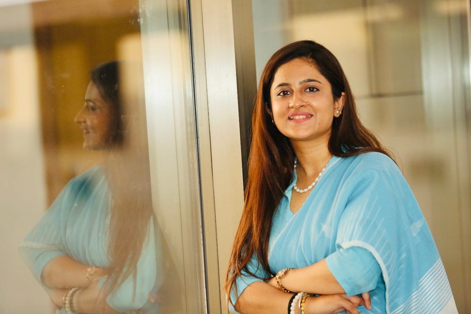 Standing with her arms folded beside a full-height glass partition that carries her reflection, in a pale blue sari.
