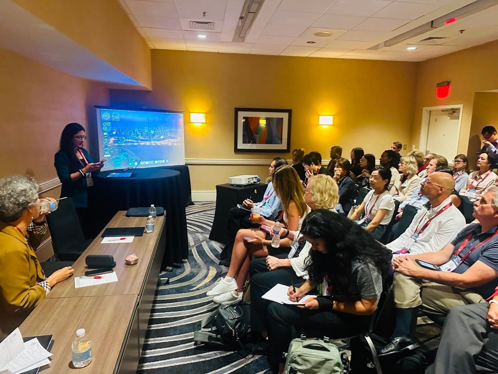 Presenting beside a large screen at the front of a conference room to an audience seated in rows, several of them taking notes.