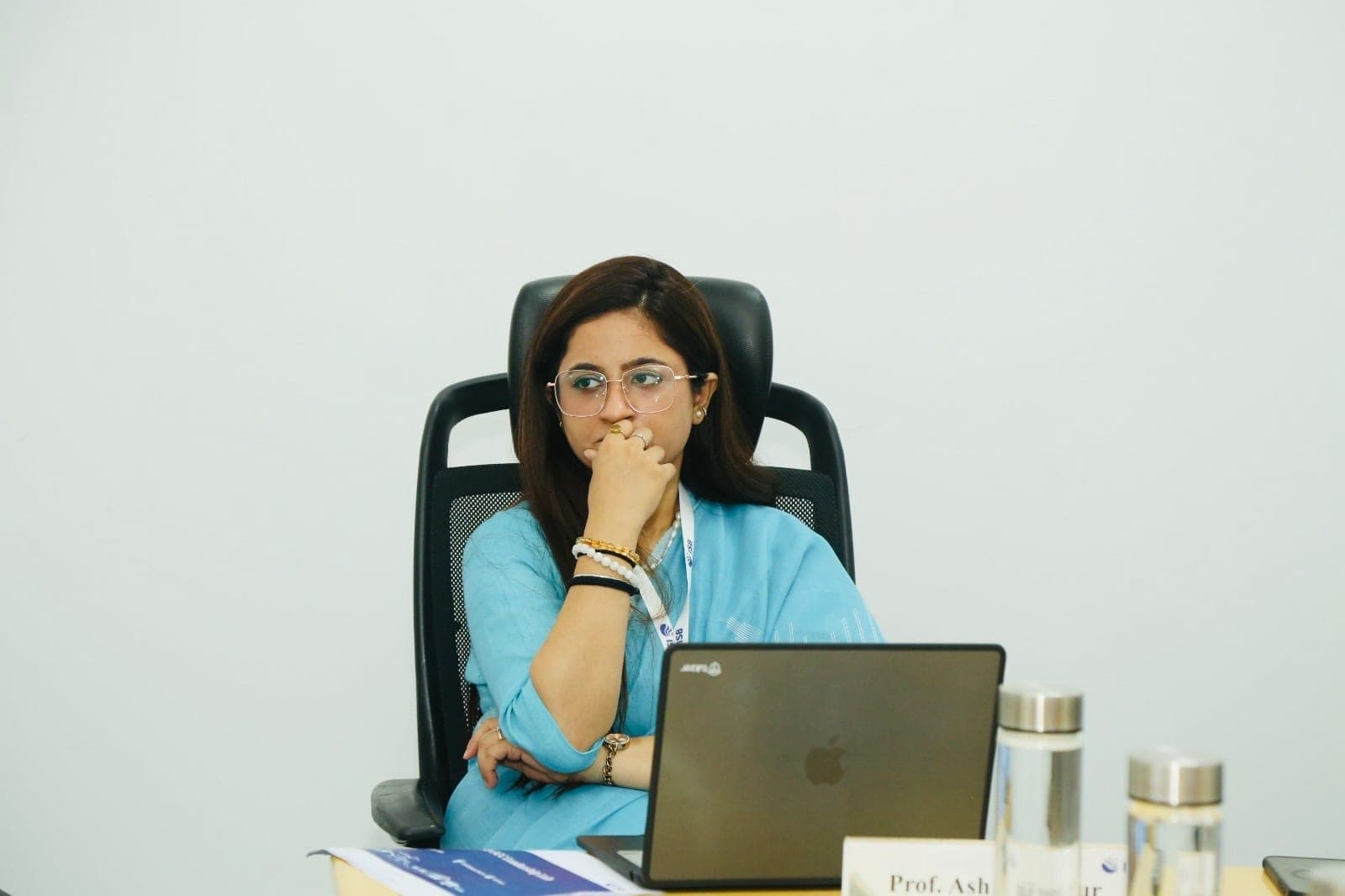 Working at a desk in a high-backed chair with an open laptop in front of her, one hand at her chin, reading from the screen.